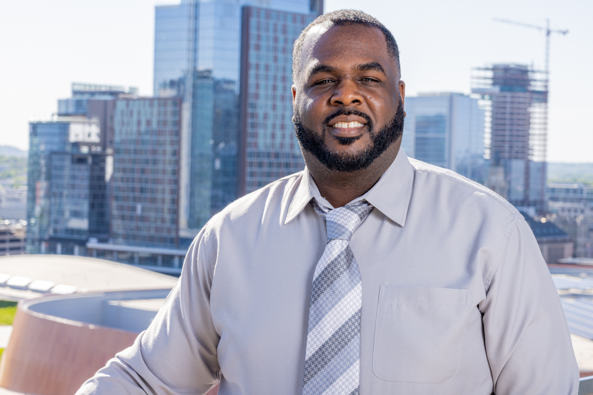 Shantheis Moody wears a gray shirt and gray striped tie. He is standing in front of a skyline. 
